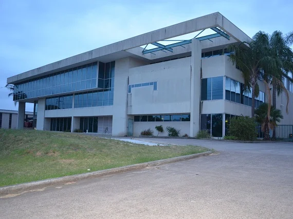 A large, modern, two-story building with glass windows and concrete structure, surrounded by a driveway and some greenery, including palm trees, under a cloudy sky.