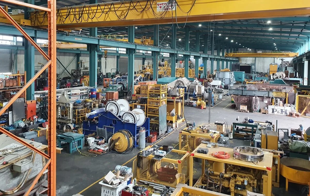 Large industrial factory interior with various machines, equipment, and metal parts scattered around. Overhead cranes and steel beams run along the ceiling, and workstations are set up throughout the spacious, well-lit area.