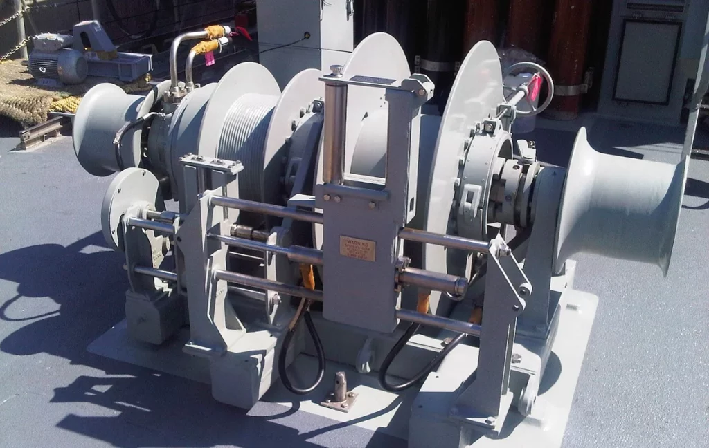 A large industrial winch with multiple drums and cables is mounted on a ship’s deck, used for pulling or hoisting heavy loads. The equipment is painted gray and surrounded by metal safety bars.