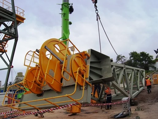 A construction crane component being lifted by another crane, while workers in safety gear guide the process at a construction site surrounded by caution tape.