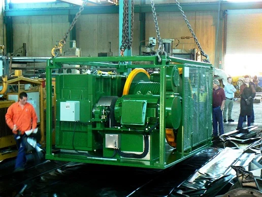 A large green industrial machine is being lifted by chains in a factory setting while several workers stand nearby observing the process.