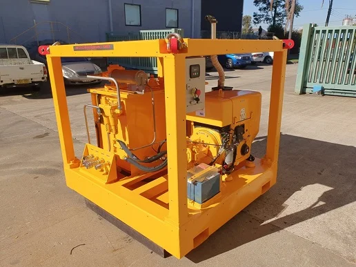 A large yellow industrial hydraulic power unit with various controls, pipes, and components, enclosed in a metal frame, is positioned outdoors on a concrete surface near parked vehicles and a building.