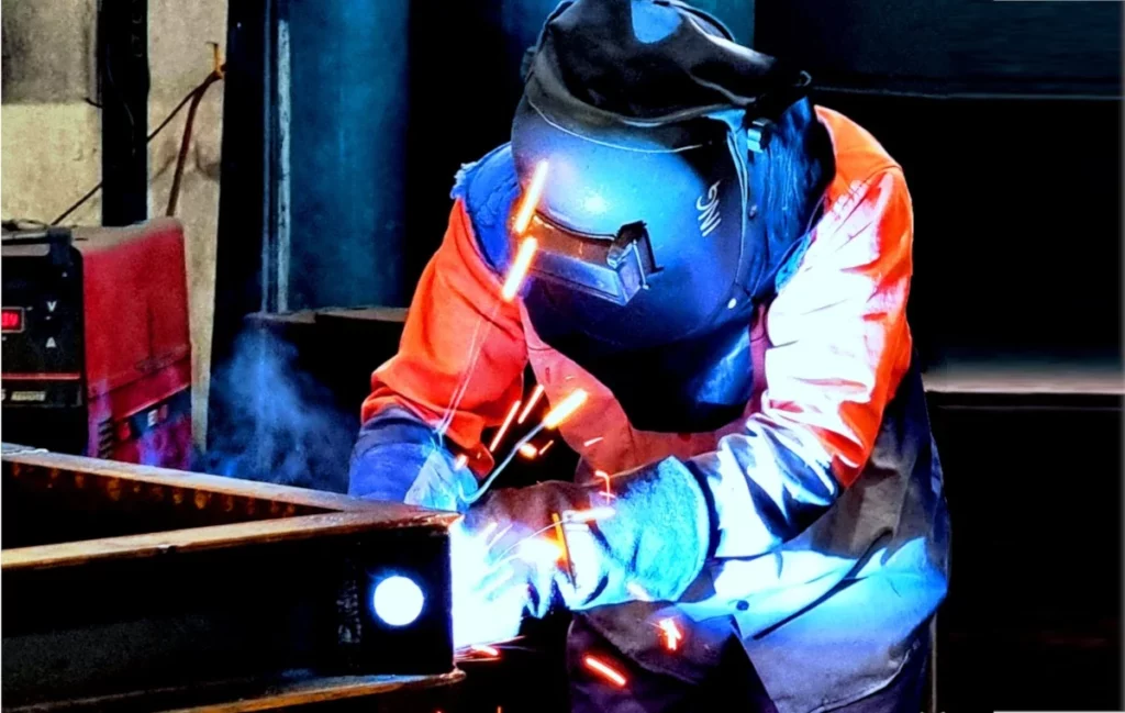A person wearing protective gear, including a welding helmet and gloves, is welding metal, with bright sparks and blue light illuminating the work area.