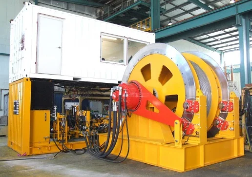 Large industrial winch system with yellow and orange components, thick steel cables wound on a drum, and a white control cabin mounted above yellow machinery inside a spacious, steel-structured industrial facility.