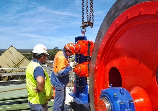 Two workers wearing safety gear and helmets inspect and work on industrial machinery outdoors. One worker is operating equipment while the other observes. Large orange and blue machine parts are visible in the foreground.