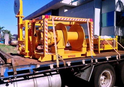 A large yellow industrial winch is secured on the back of a flatbed truck, parked outside a building. The winch is surrounded by safety barriers and labeled “WP020.” Other equipment is visible beside it.