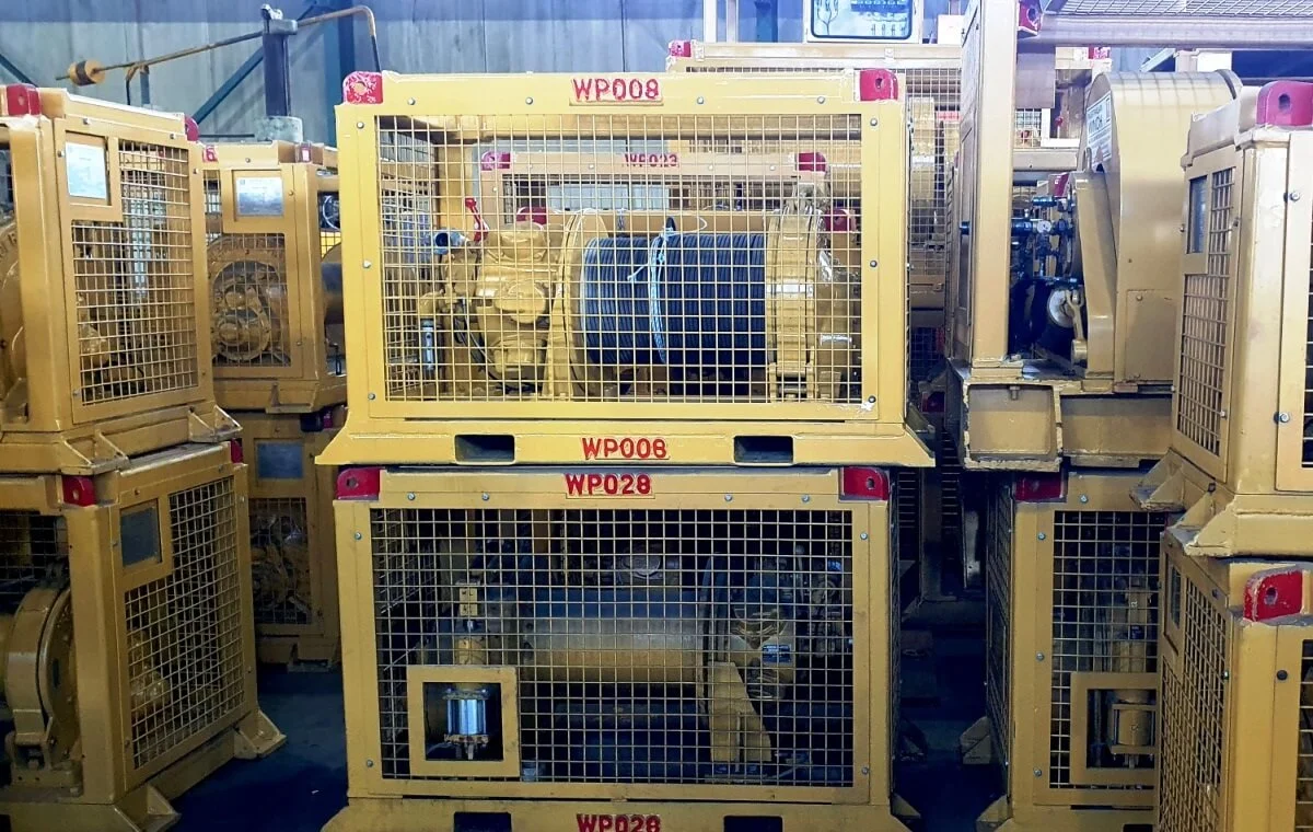 Several large, yellow industrial machines are enclosed in metal cages and stacked in a warehouse. Each cage is labeled with a red identification number. The machines appear to be organized and stored for use.