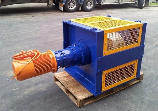A blue and yellow industrial winch with an orange motor, mounted on a wooden pallet, sits on concrete outdoors near a large truck.