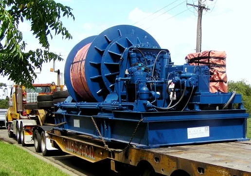A large blue industrial winch with a thick cable spool sits on a flatbed trailer, ready for transport. The equipment is secured, and a utility pole and trees are visible in the background.